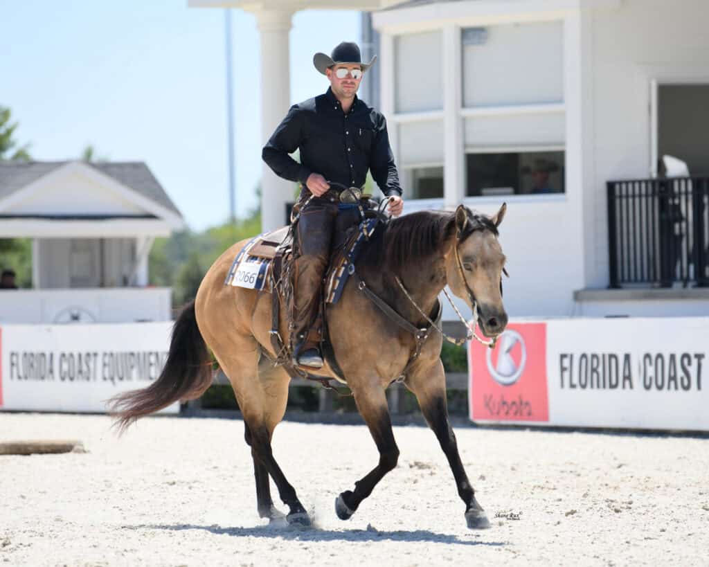 A man in a black shirt and cowboy hat rides a brown horse in an outdoor arena near a white building.