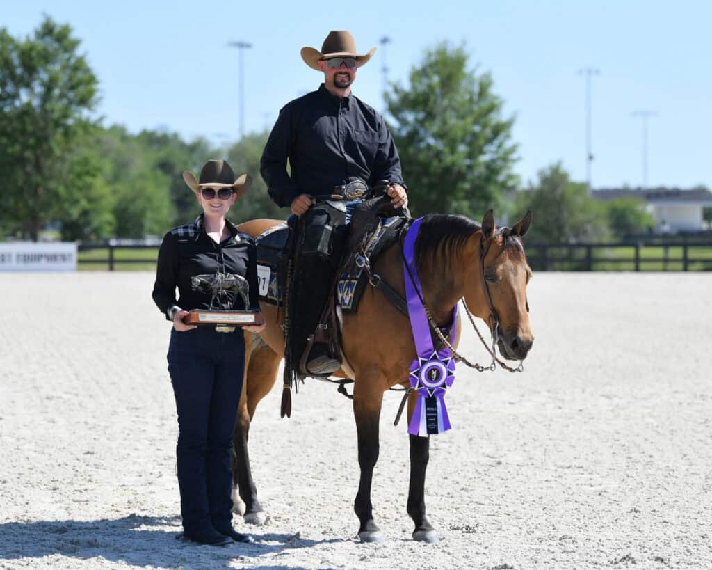A man in a cowboy hat sits on a horse with a purple ribbon; a woman in a cowboy hat stands beside them holding a trophy. They are outdoors on a sandy arena.