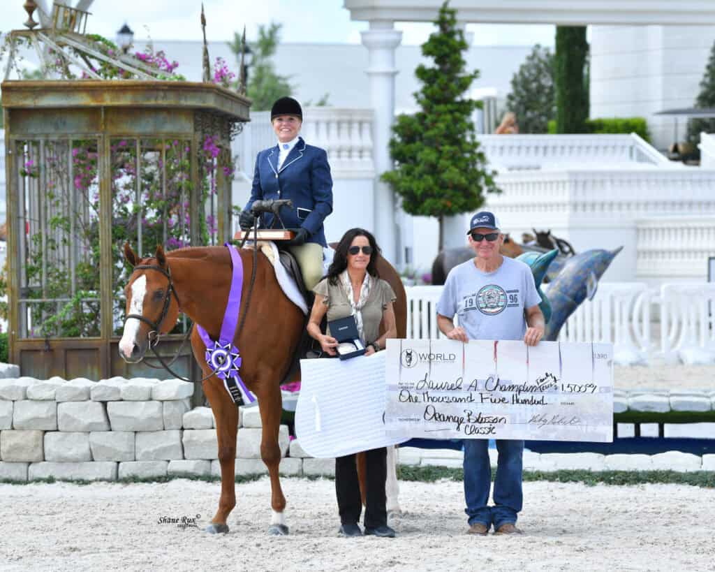 A rider on a horse with a purple ribbon, flanked by two people holding a trophy and a large check, stands in an outdoor arena.