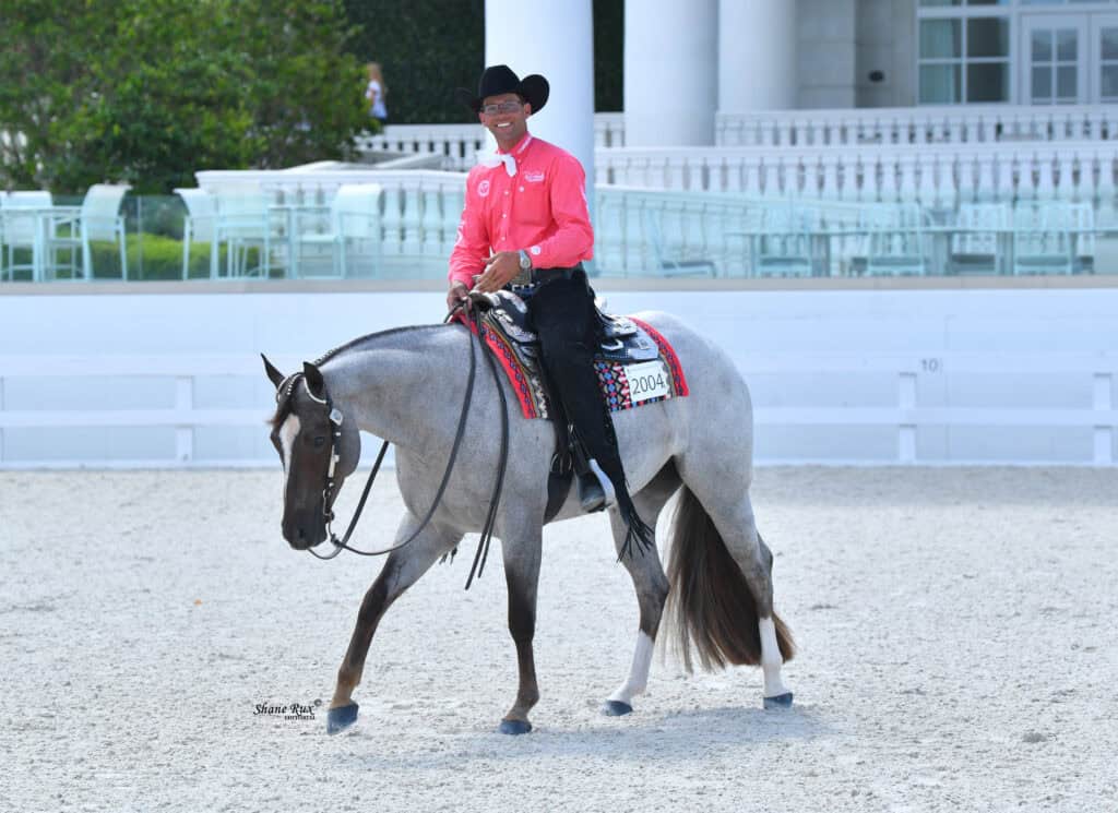 A man in a bright pink shirt and black cowboy hat rides a gray horse with a decorative saddle pad in an outdoor arena.