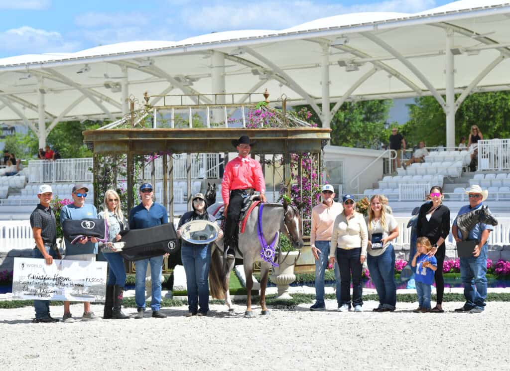 A group of people pose with a horse and rider, holding awards and a large check, in an outdoor arena with white seating and decorative flowers in the background.