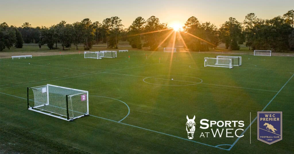 A large outdoor soccer field complex with multiple goalposts, set on green grass at sunrise. Logos for 
