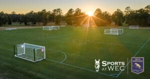 A large outdoor soccer field complex with multiple goalposts, set on green grass at sunrise. Logos for 