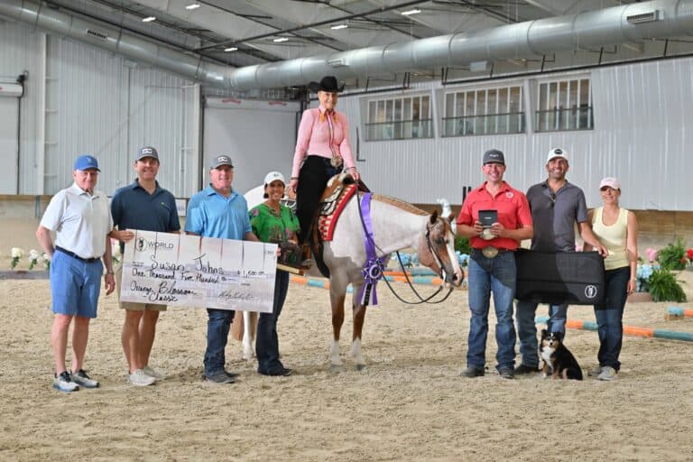 A group of people stands in an indoor arena with a horse and rider; one person holds a large check and another holds a trophy, with a dog sitting in front of the group.