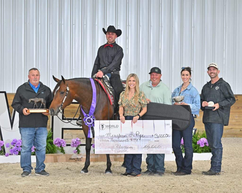 A group of six people poses indoors with a horse, awards, and a large check for $3,000 at a horse show event decorated with purple flowers.
