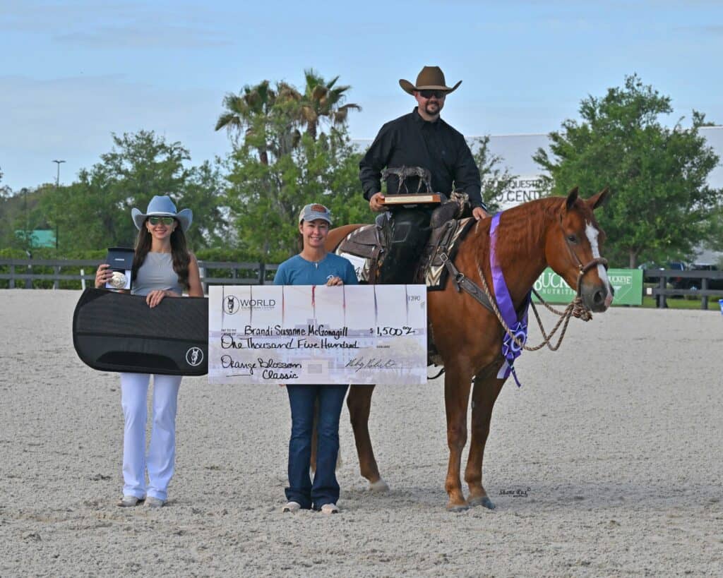 Three people pose with a winner's check and prizes; one is on horseback wearing a cowboy hat and holding a trophy, the others stand beside the horse holding a check and a guitar case.