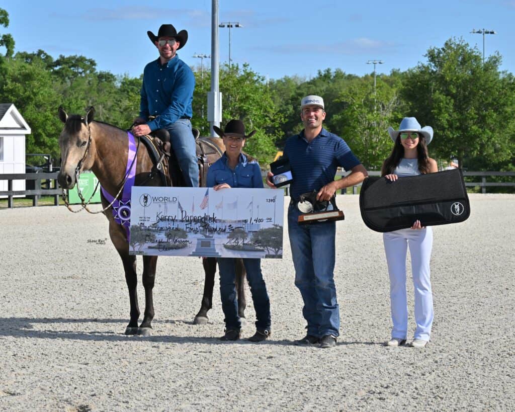 Four people pose outdoors with a horse, a large check, a trophy, and an award case. One person is mounted on the horse; the others stand on the ground, all wearing hats and casual attire.