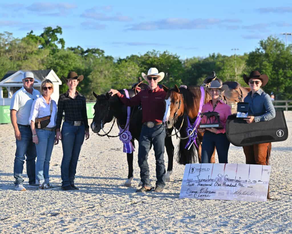 A group of six people stands with two horses, award ribbons, a large check, and a guitar case at an outdoor equestrian event.