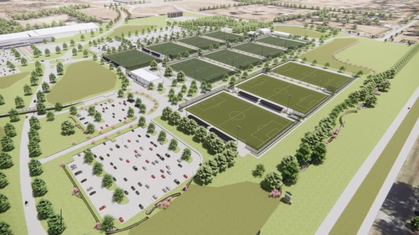 Aerial view of a large sports complex with multiple soccer fields, surrounding trees, nearby buildings, and a parking lot filled with cars.