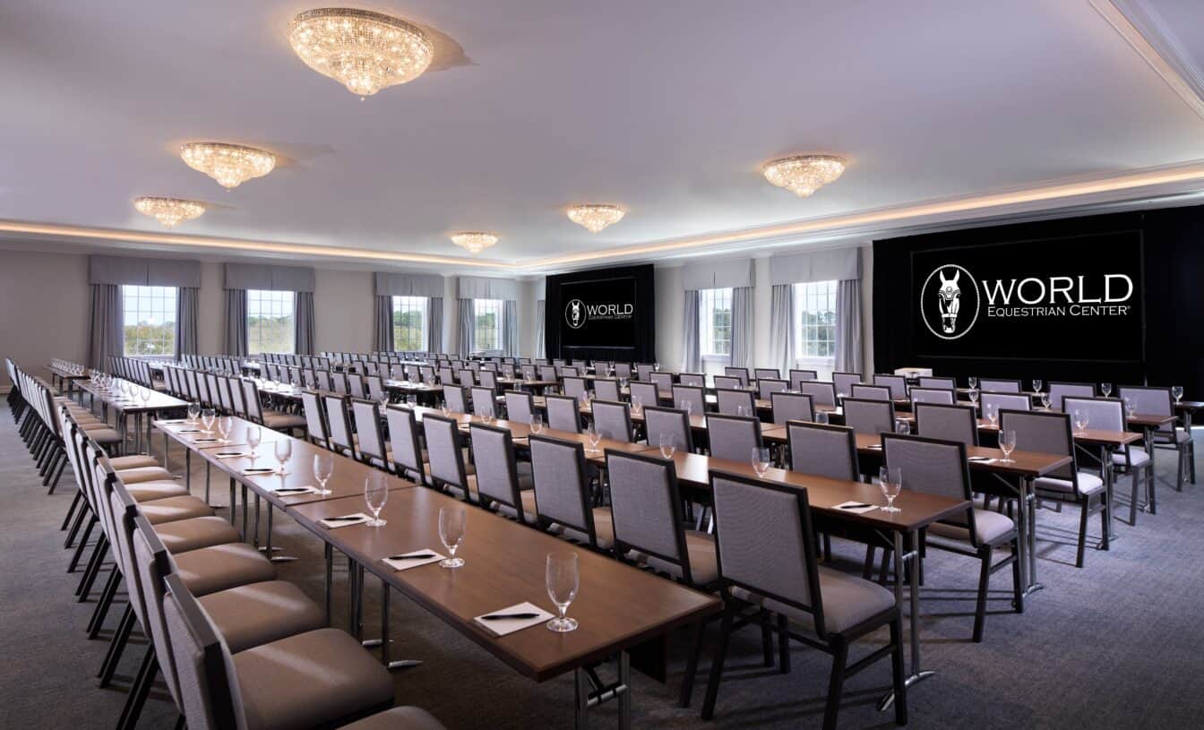 A conference room with rows of tables and chairs, empty glasses and notepads, and two large screens displaying the World Equestrian Center logo.