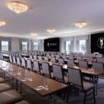 A conference room with rows of tables and chairs, empty glasses and notepads, and two large screens displaying the World Equestrian Center logo.