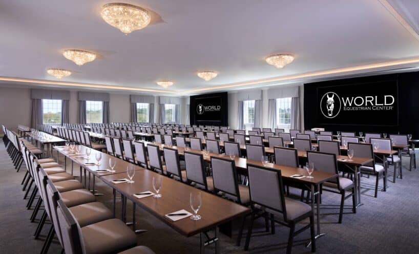 A conference room with rows of tables and chairs, empty glasses and notepads, and two large screens displaying the World Equestrian Center logo.