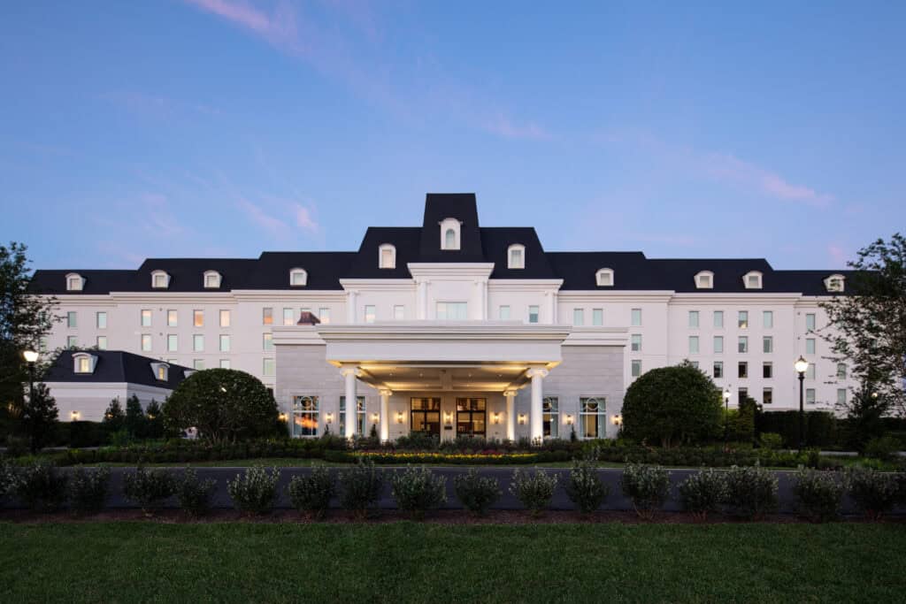 A large, white, multi-story hotel building with black roof and dormer windows, viewed from the front at dusk with a landscaped entrance and driveway.