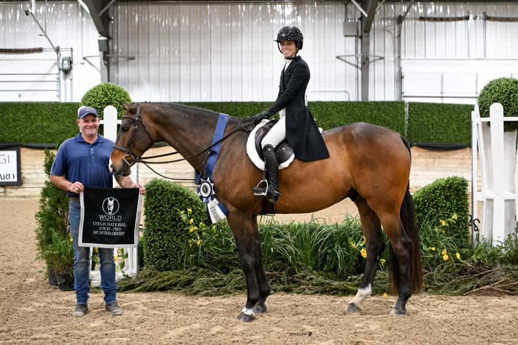 A rider on a horse wearing a blue ribbon poses next to a smiling man holding a World Horse Show finalist banner in an indoor arena.