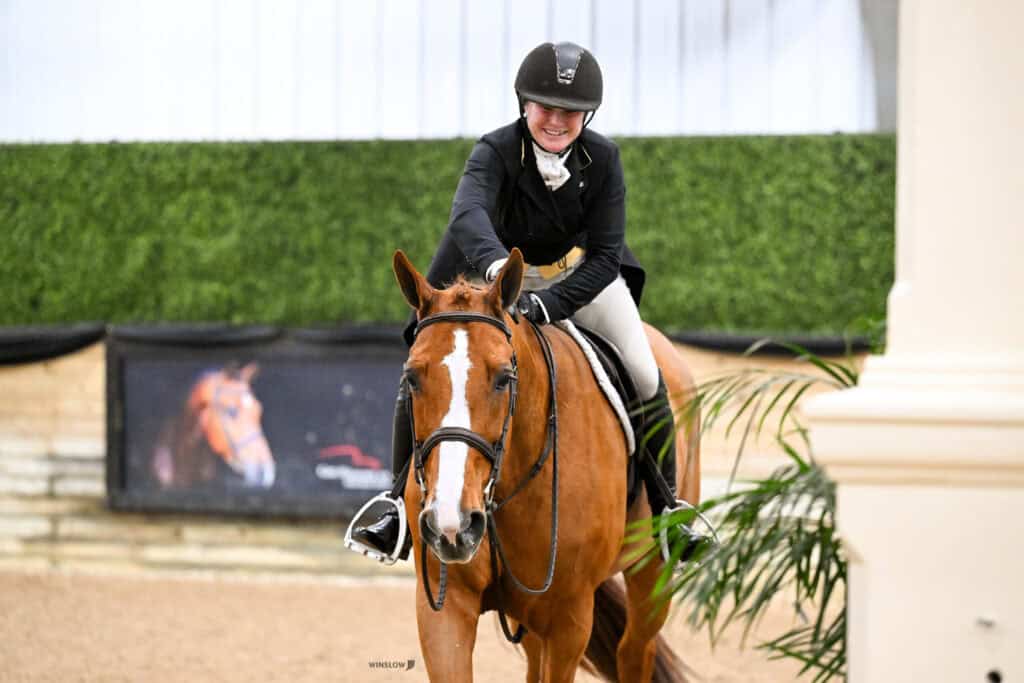 A rider in equestrian attire smiles while sitting on a brown horse in an indoor arena with green hedge walls and a horse portrait in the background.