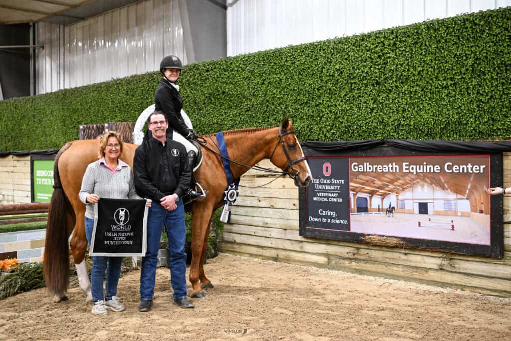 A rider on horseback poses indoors with two people standing beside them, in front of a banner for the Galbreath Equine Center at Ohio State University.