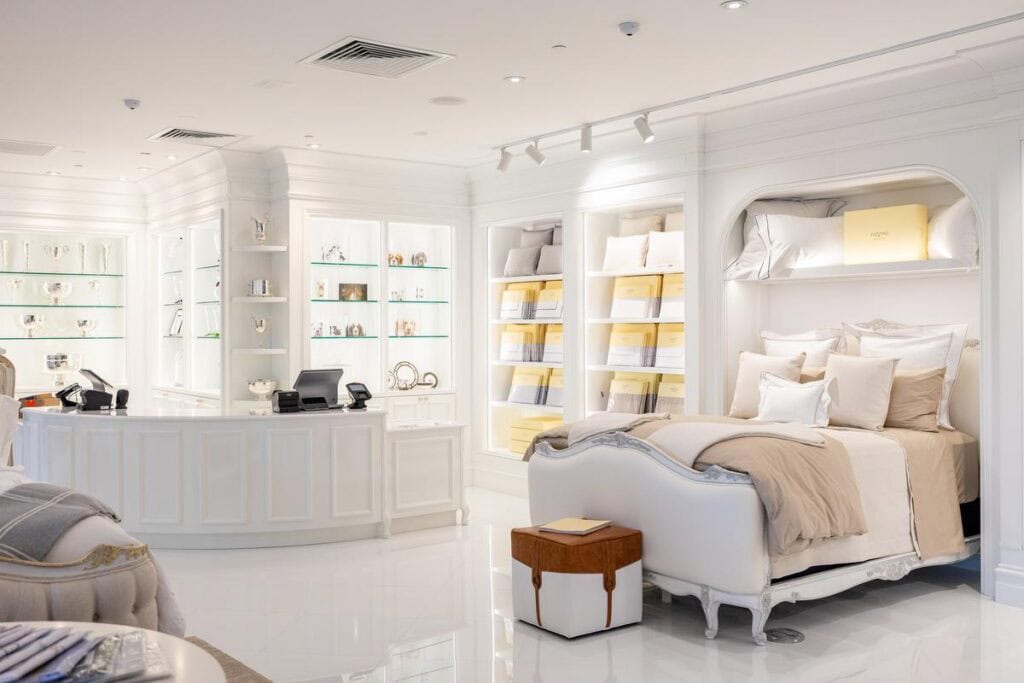 A bright, upscale store interior with a white and beige bed display, shelves with linens, glass cabinets, and a curved checkout counter.