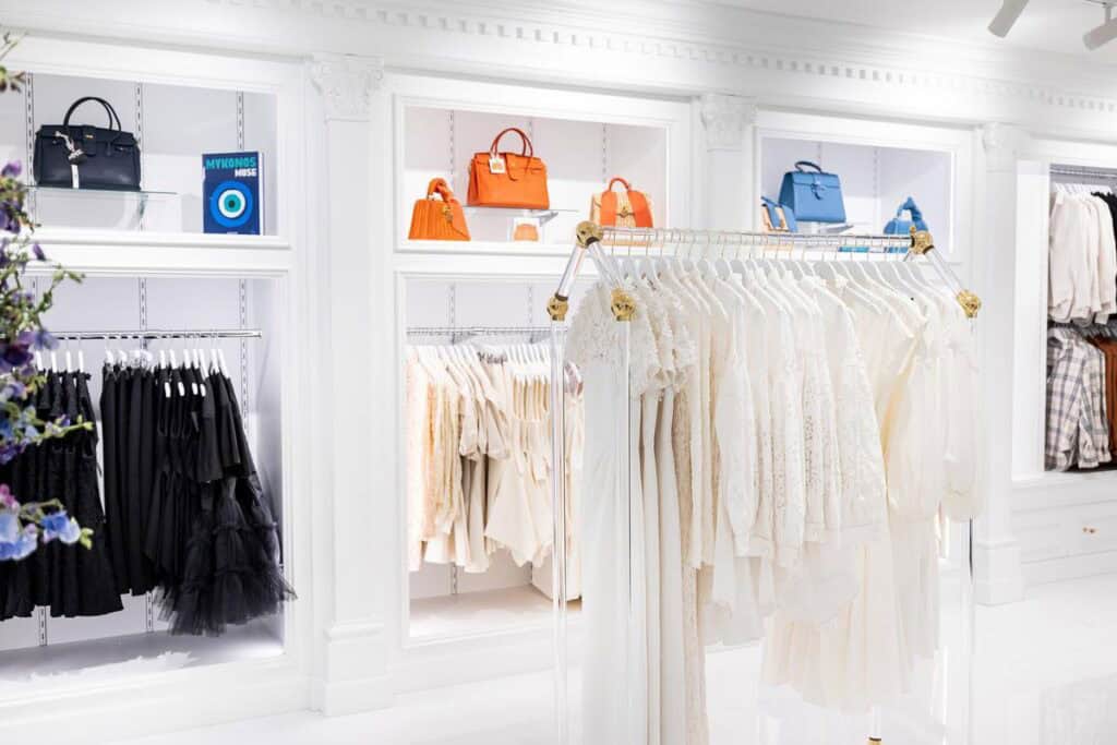 A clothing store displays white and beige garments on a rack, with colorful handbags and folded clothes arranged on white shelves in the background.