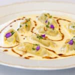 A plate of stuffed dumplings in a creamy white sauce, garnished with edible flowers, pine nuts, chopped herbs, and a drizzle of brown sauce.