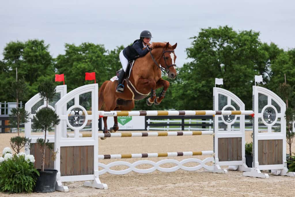 A rider wearing a helmet jumps a brown horse over a high obstacle during an equestrian event in an outdoor arena.