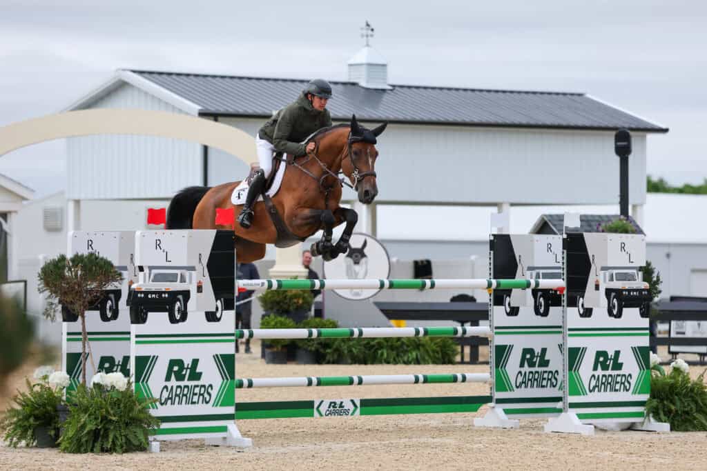 A rider on a brown horse jumps over a green and white obstacle during an equestrian show jumping event, with a barn and sponsor signs in the background.