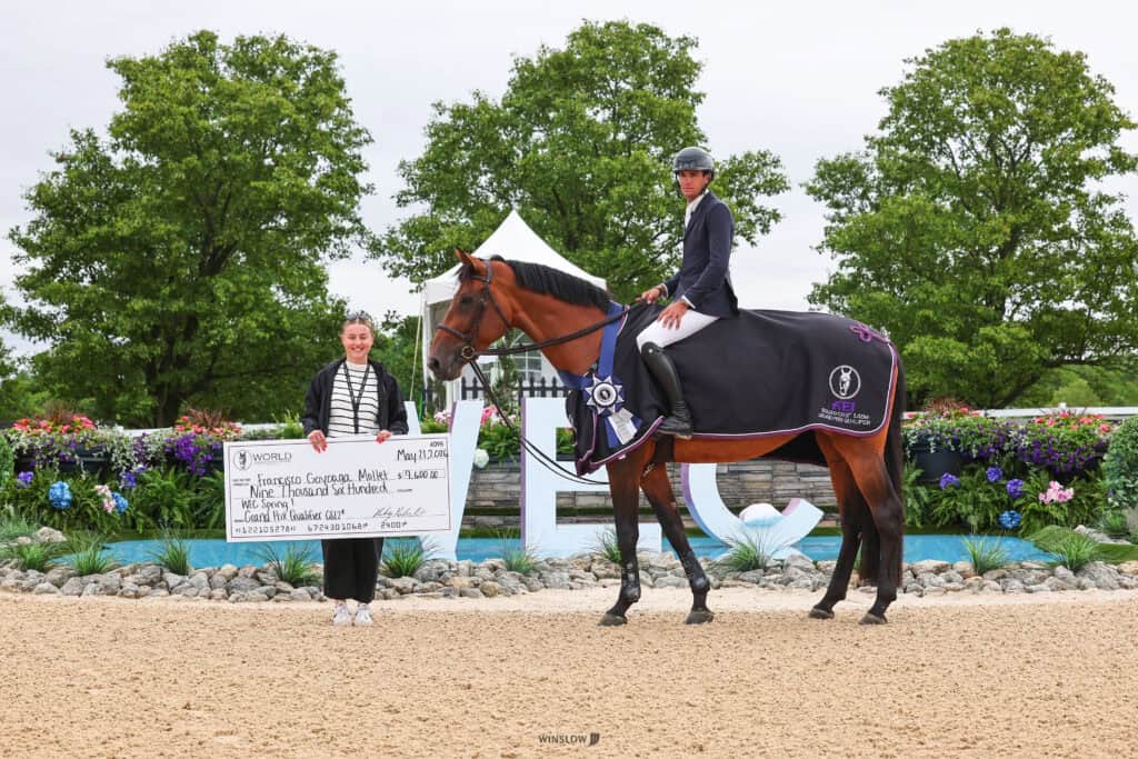 A woman holds a large check while a man in riding gear sits on a horse, both posing on a gravel arena with greenery and flowers in the background.