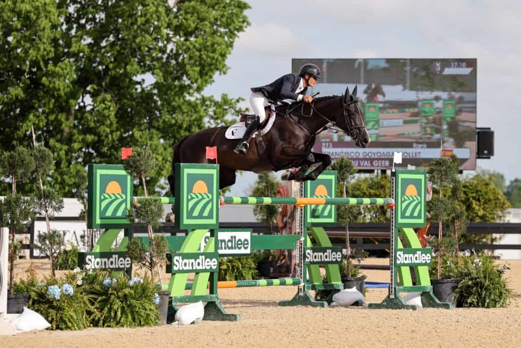 Equestrian rider and horse leap over a green and yellow obstacle during a show jumping competition, with a scoreboard visible in the background.