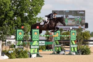 Equestrian rider and horse leap over a green and yellow obstacle during a show jumping competition, with a scoreboard visible in the background.