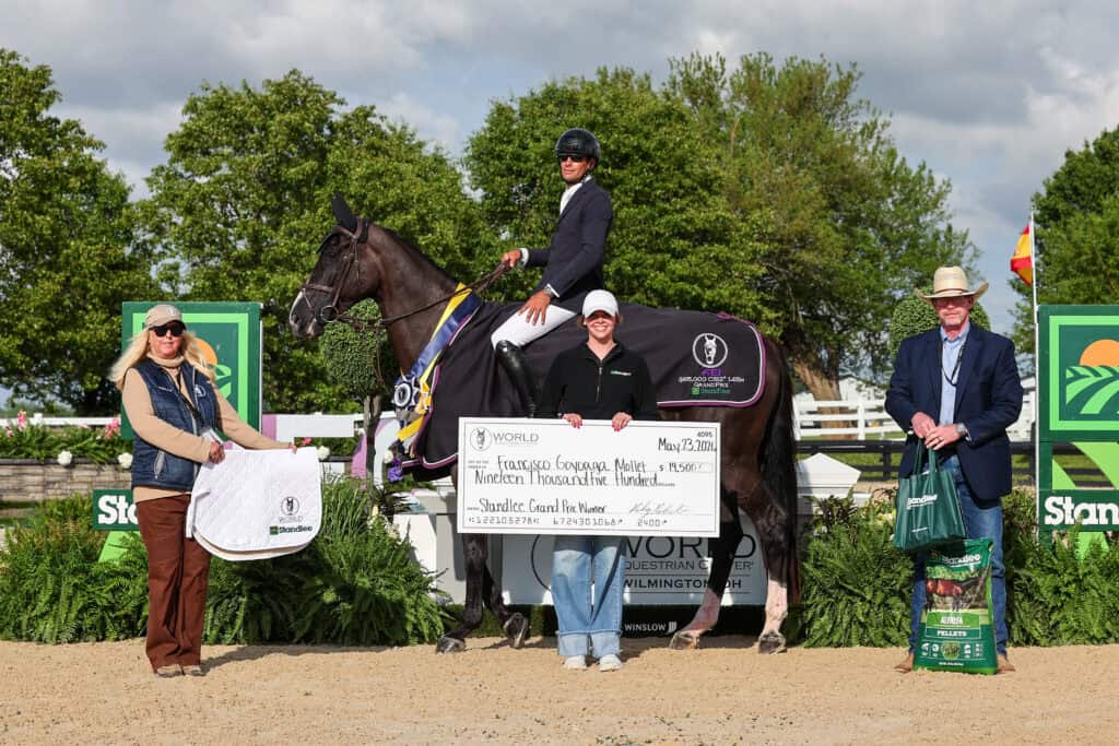 Four people pose outdoors with a horse and an oversized check at an equestrian event. One person sits on the horse, while the others stand beside them, smiling at the camera.