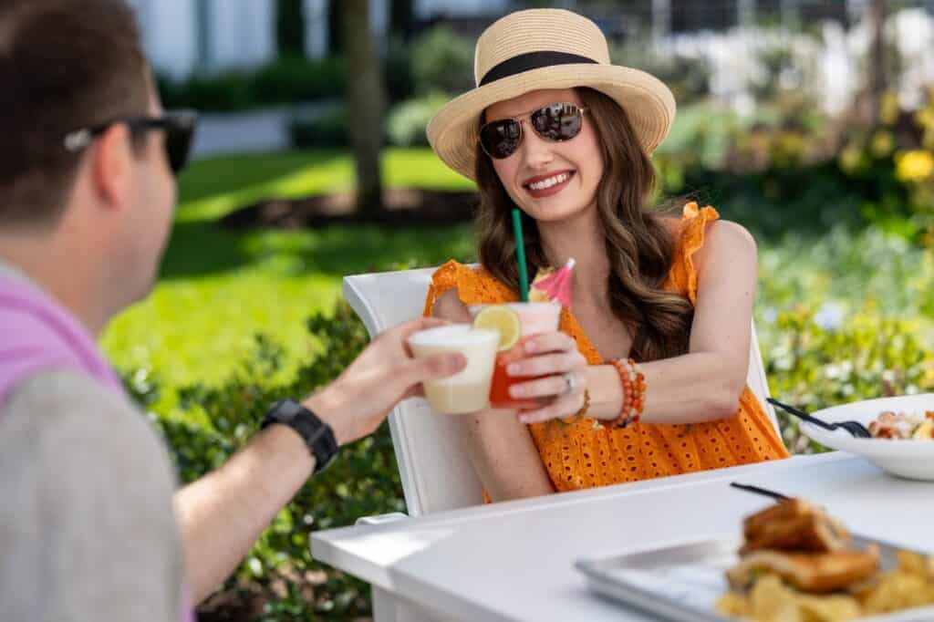 Two people sit at an outdoor table, toasting with drinks. One person wears a hat and sunglasses, smiling and holding a pink cocktail with a straw. Food is visible on the table.