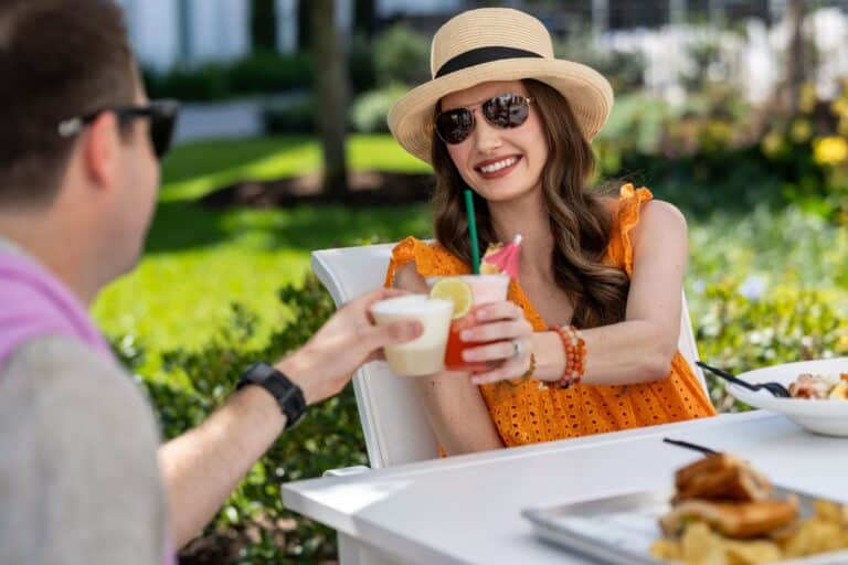 Two people sit at an outdoor table, toasting with drinks. One person wears a hat and sunglasses, smiling and holding a pink cocktail with a straw. Food is visible on the table.