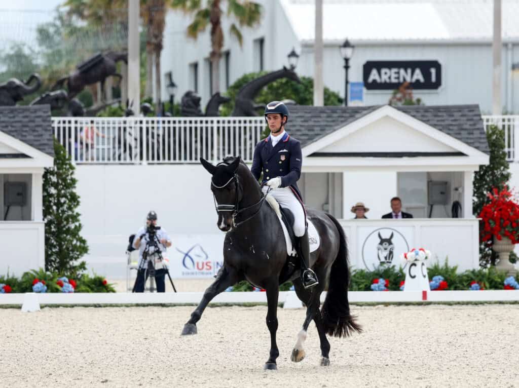 Equestrian rider in formal attire performs dressage on a black horse in an outdoor arena marked "Arena 1," with cameras and spectators in the background.