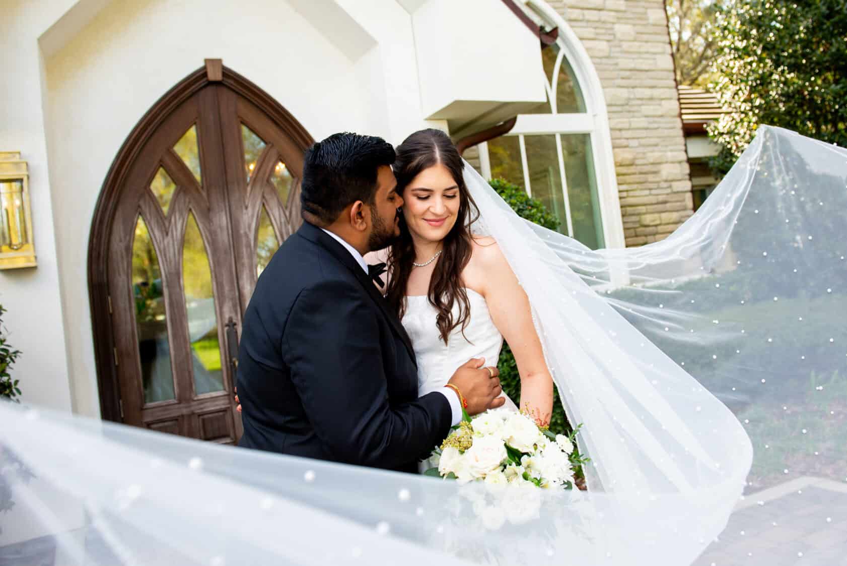 A groom kisses the bride on the cheek as they stand outside a building; the bride holds a bouquet and her veil is spread out around them.