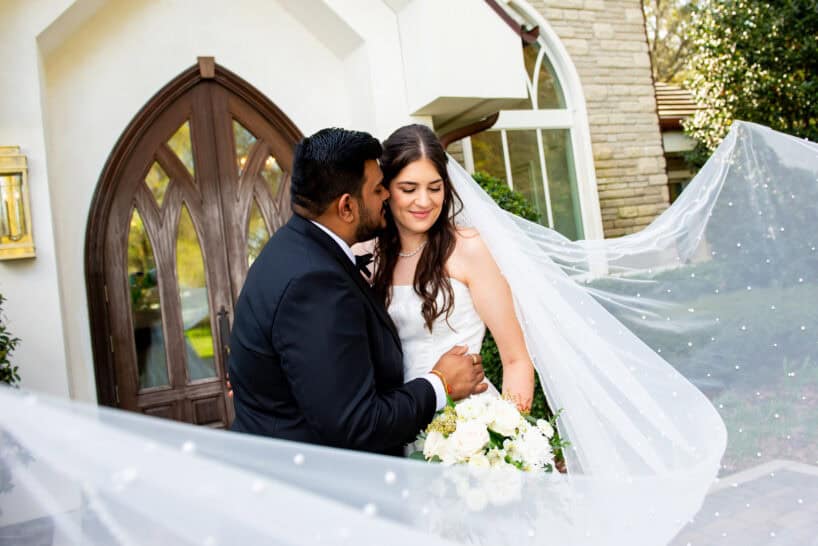 A groom kisses the bride on the cheek as they stand outside a building; the bride holds a bouquet and her veil is spread out around them.