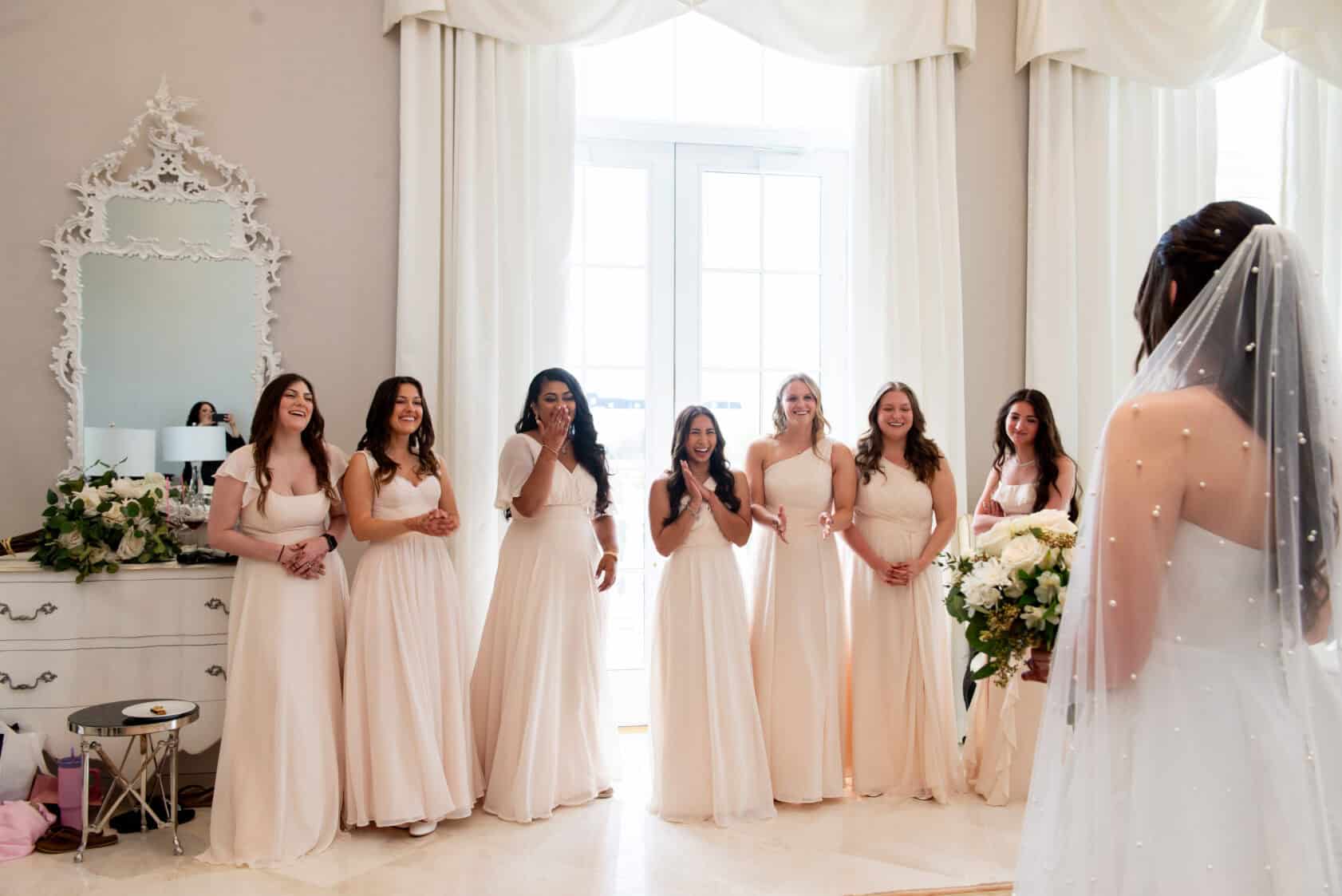 A bride in a white dress and veil stands holding a bouquet, facing six bridesmaids in matching light pink dresses inside a bright, elegant room.