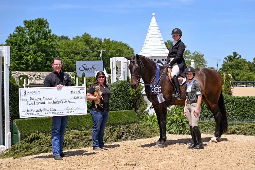 Four people pose outdoors with a large horse; one person is mounted, two hold a large check, and one holds a ribbon. The background has greenery and a white tower.