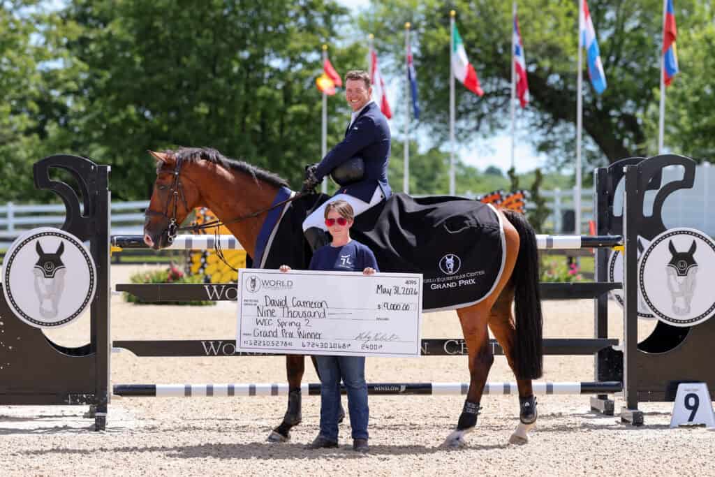 A man on horseback holds a winning ribbon, while another person in sunglasses holds a large check for $9,000 in front of a jumping arena with international flags in the background.