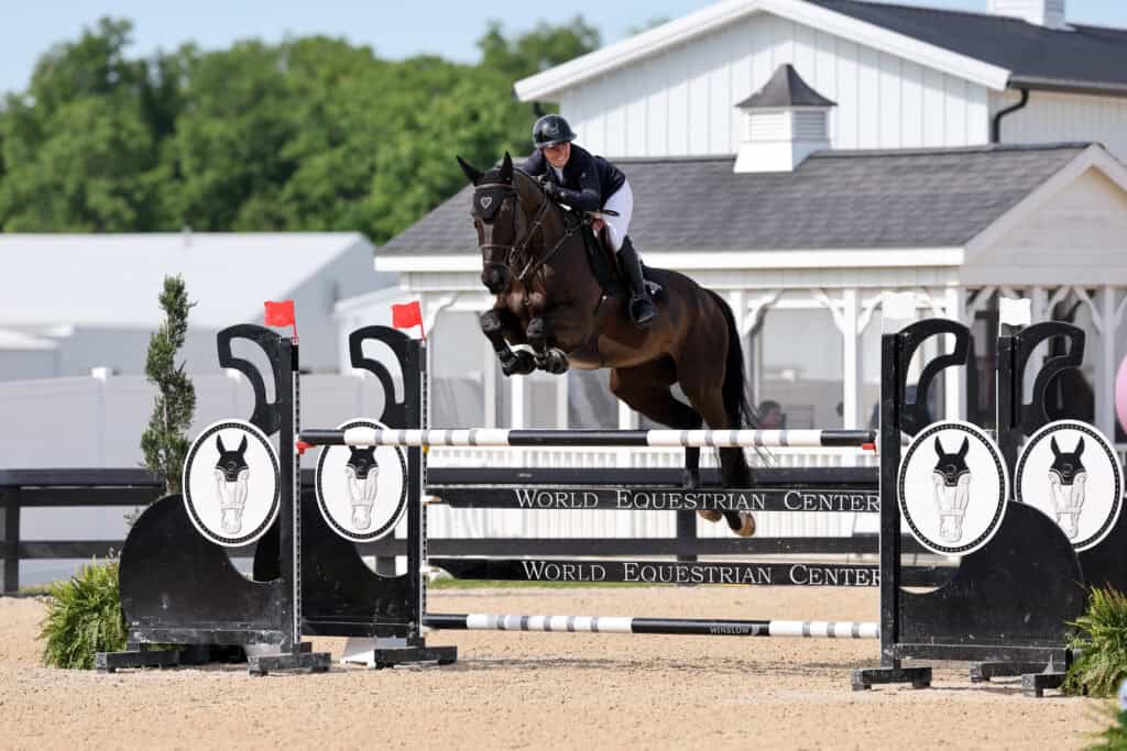 A rider and horse jump over an obstacle in an outdoor equestrian arena, with the World Equestrian Center logo visible on the jump.