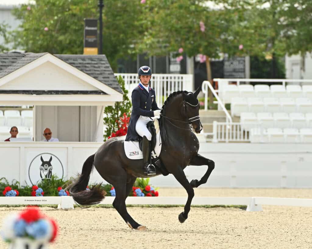 A rider in formal attire performs dressage on a dark horse in an outdoor arena with white fences and empty seating in the background.