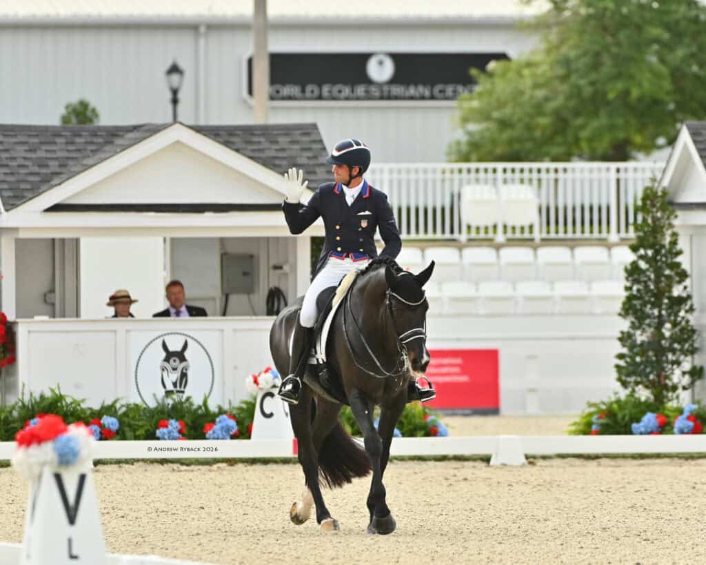 A dressage rider in formal attire waves while riding a black horse in an outdoor arena, with a 