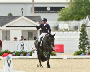 A dressage rider in formal attire waves while riding a black horse in an outdoor arena, with a 