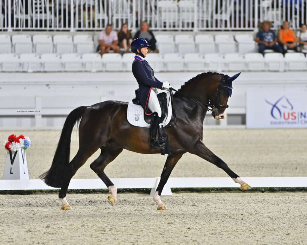 A rider in formal equestrian attire guides a dark horse performing a dressage movement in an arena with white stands and a US dressage sign in the background.