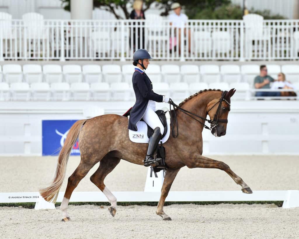 A rider wearing a helmet and formal attire guides a horse in an extended trot during a dressage competition in an outdoor arena.