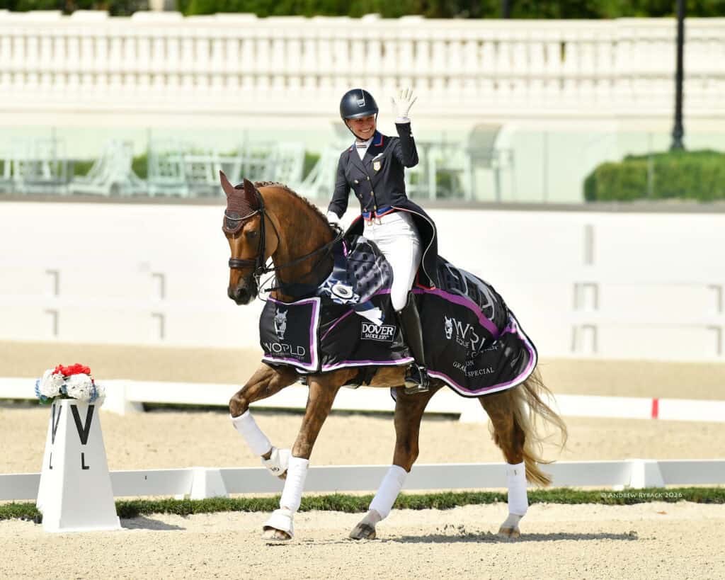 Equestrian rider in formal attire waves while riding a horse draped in a branded blanket during a dressage competition in an outdoor arena.