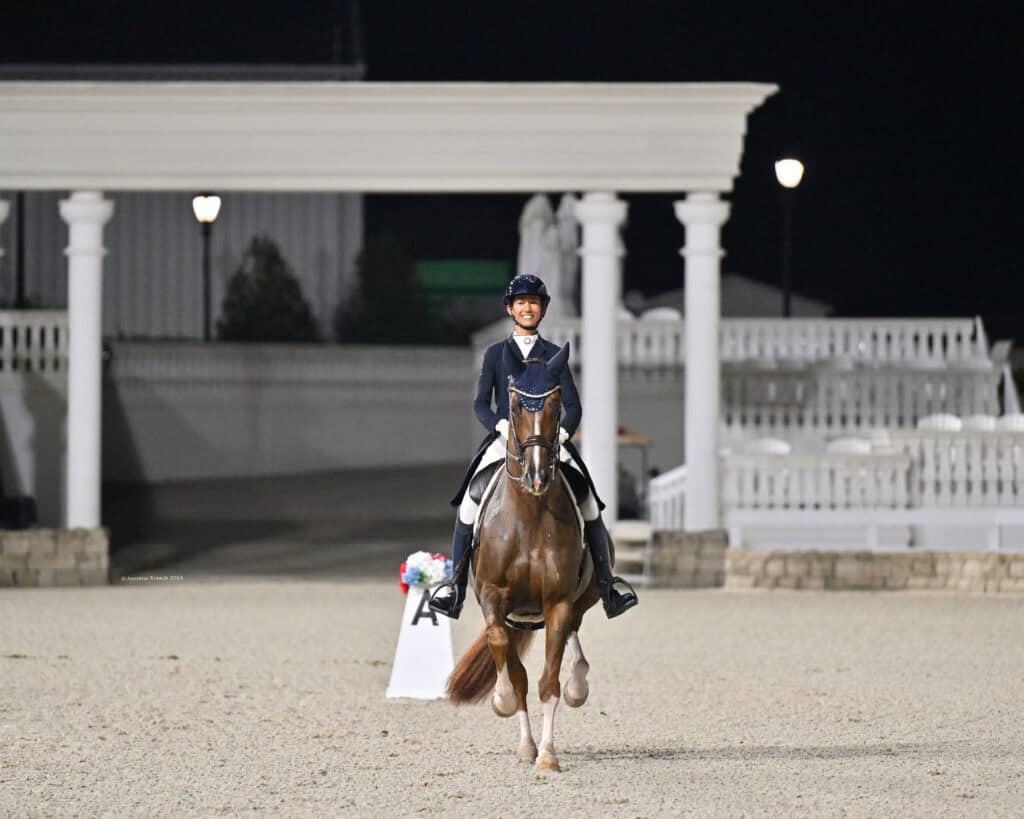 A rider in equestrian attire sits atop a brown horse, performing in a dressage arena at night with white columns and empty seating in the background.