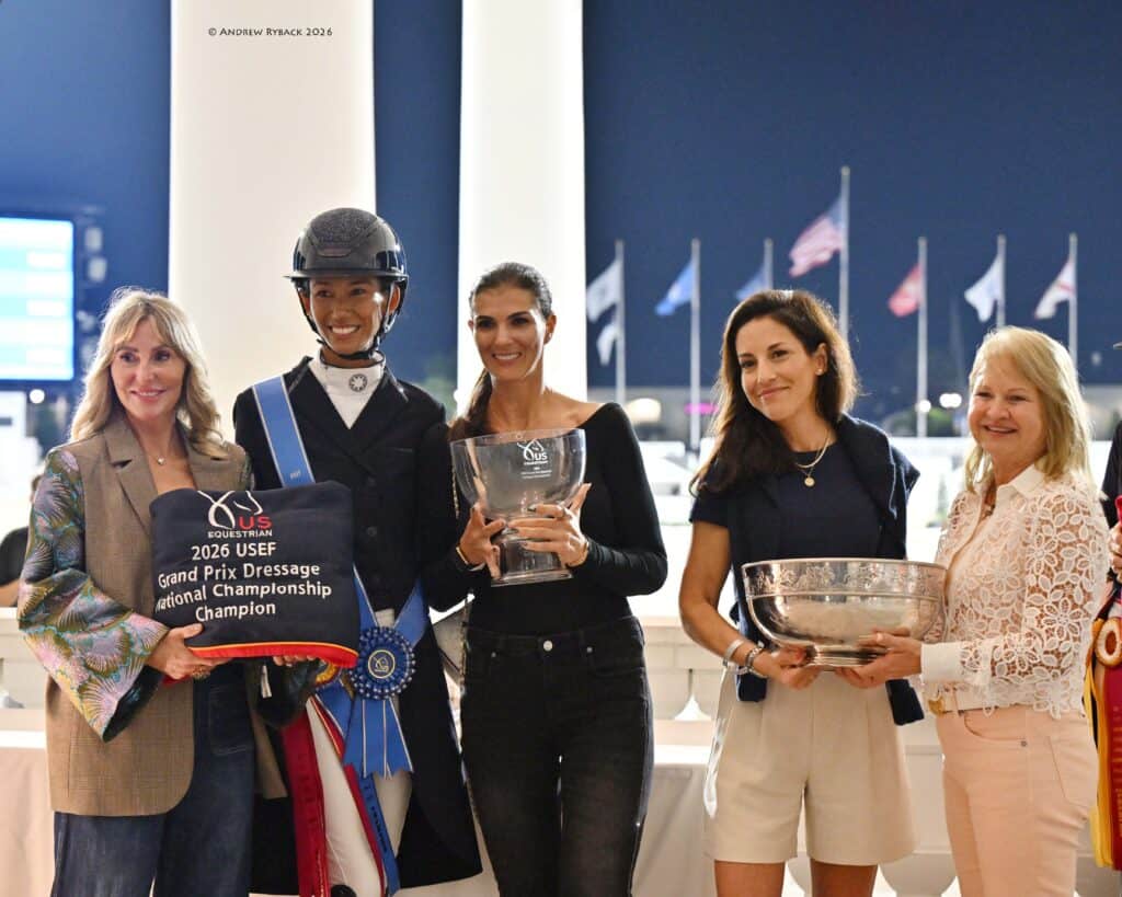 Five women stand in a row at an awards ceremony; one holds a ribbon and championship sash, while others hold trophies and a plaque. Flags are visible in the background.