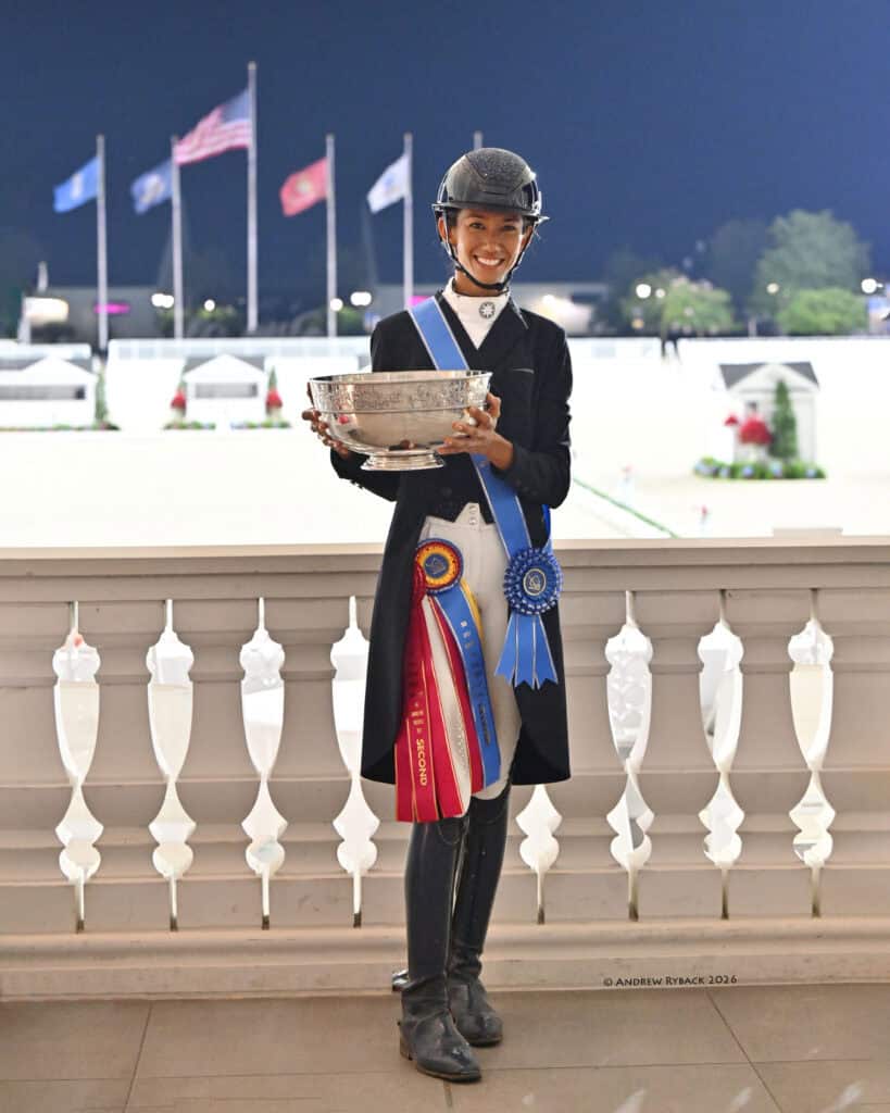 A smiling equestrian dressed in riding attire stands on a balcony holding a large trophy, with blue and red award ribbons pinned to their outfit. Four flags wave in the background.