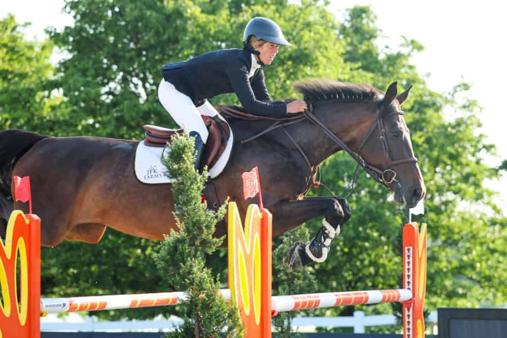 A person wearing a helmet and riding attire is jumping a horse over a red and yellow obstacle during an equestrian event, with green trees in the background.