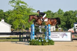 A rider in equestrian gear guides a horse over a jump at an outdoor competition, with a Kentucky Equine Research sign visible in the background.