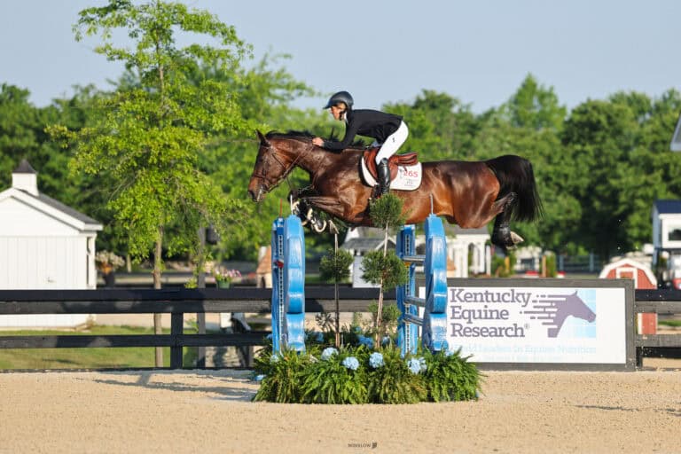 A rider in equestrian gear guides a horse over a jump at an outdoor competition, with a Kentucky Equine Research sign visible in the background.